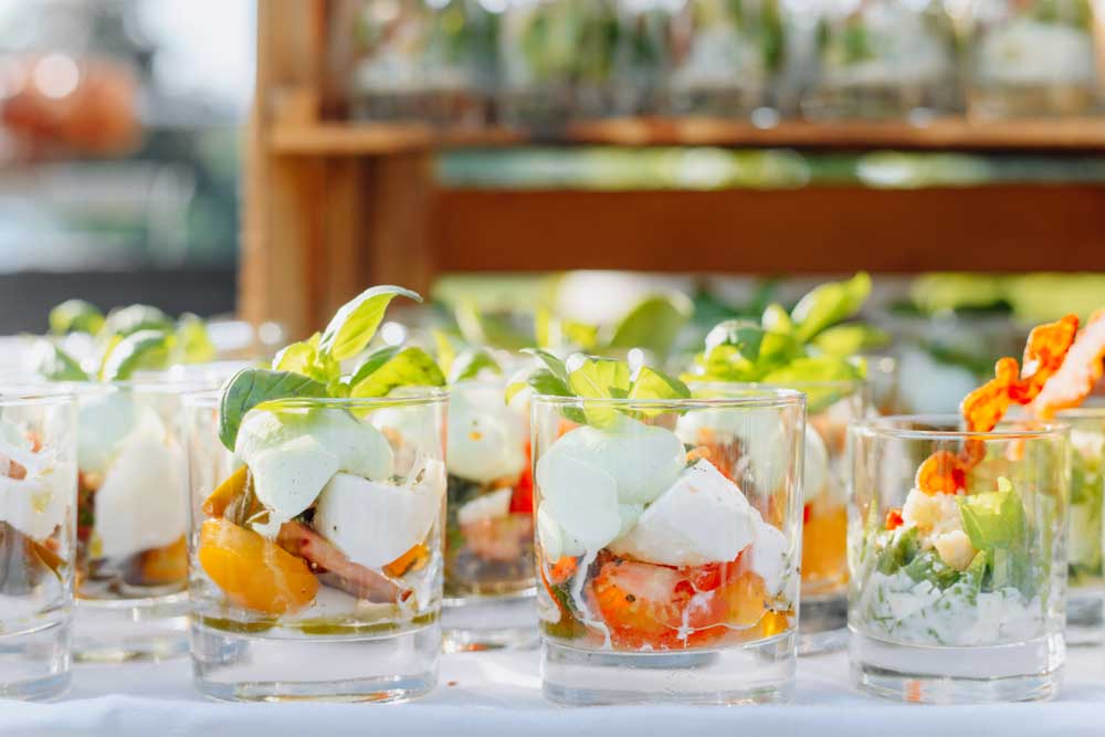 Small servings of carpese salad inside glass cups lines up on a white-clothed table